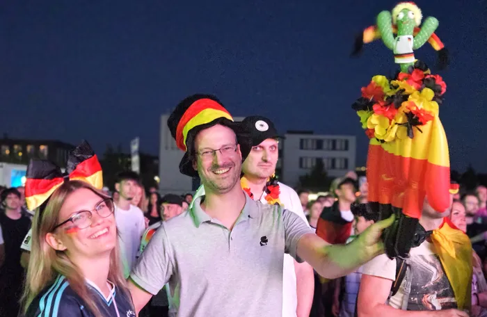 Jubelnde Fans mit Deutschlandfahnen und -hüten feiern bei einer Public-Viewing-Veranstaltung im Freien. Im Vordergrund ein lächelnder Mann, der eine Deutschlandflagge hochhält.