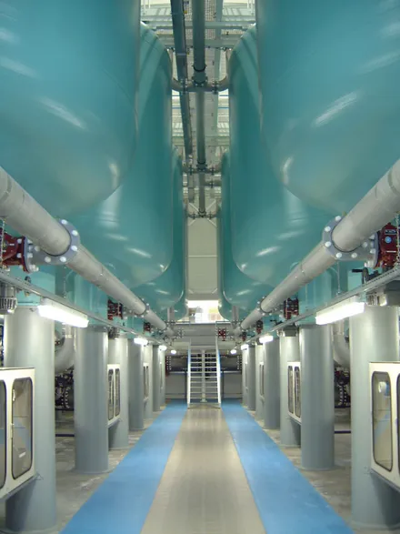 Innenansicht einer Wasseraufbereitungsanlage mit großen, türkisfarbenen Rohren an der Decke und einer Reihe von grauen Schaltschränken an den Seiten eines blauen Flurs.