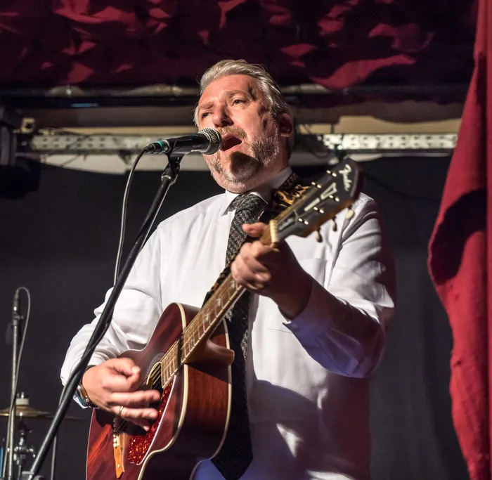Ein Musiker in weißem Hemd und Krawatte spielt Akustikgitarre und singt in ein Mikrofon beim Benefizkonzert „Rock for Peace“.