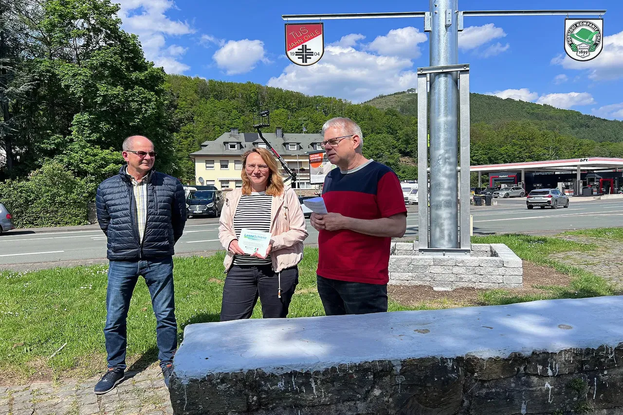 Drei Personen stehen vor einem Wappenbaum und halten Dokumente oder Notizen. Im Hintergrund ist eine Tankstelle und eine Landschaft mit Hügeln zu sehen.