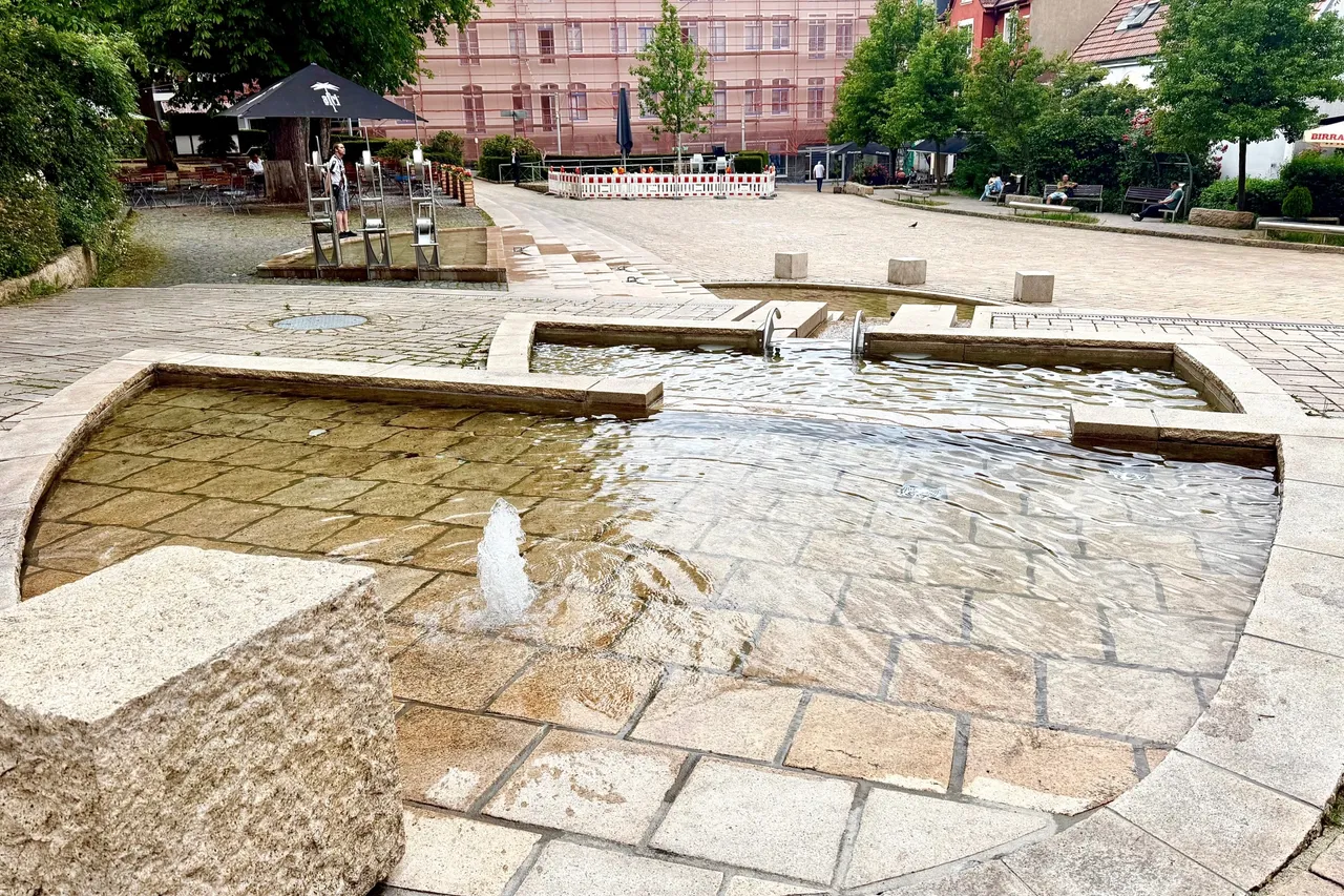 Ein rechteckiges Wasserspiel mit einem zentralen Wasserstrahl befindet sich auf einem Platz mit Pflastersteinen. Im Hintergrund sind Gebäude und einige Sitzgruppen zu sehen.