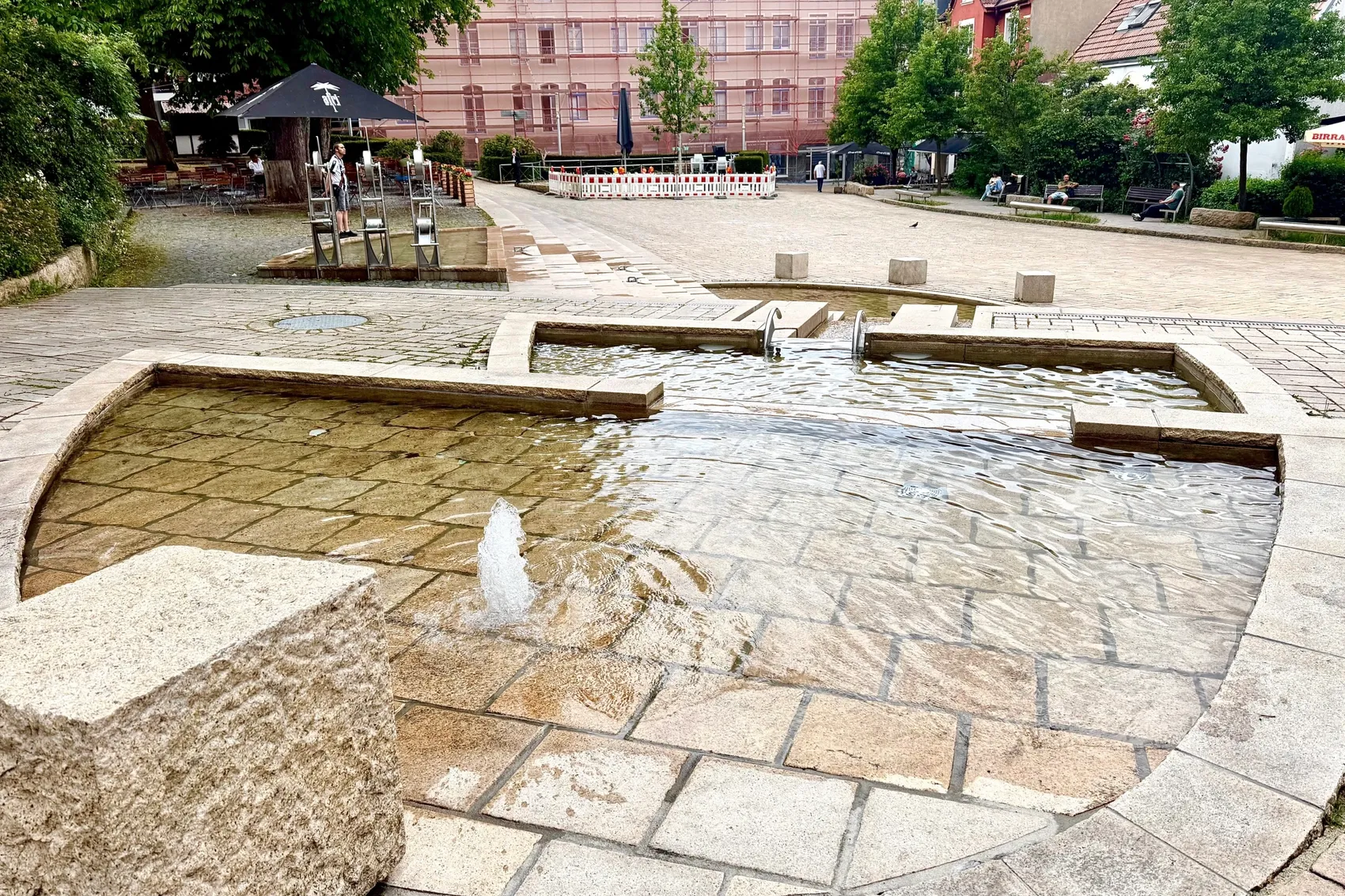 Ein rechteckiges Wasserspiel mit einem zentralen Wasserstrahl befindet sich auf einem Platz mit Pflastersteinen. Im Hintergrund sind Gebäude und einige Sitzgruppen zu sehen.