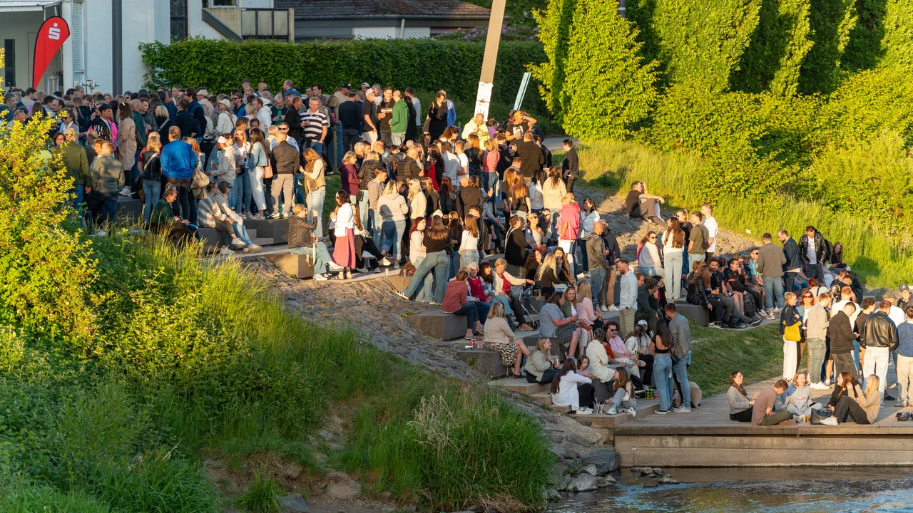Große Menschenmenge an einem sonnigen Tag an einem Flussufer, viele sitzen oder stehen auf einer Uferpromenade und Grasflächen.