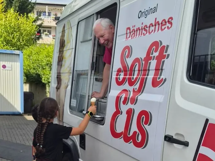 Ein lächelnder Mann im Fenster eines Softeis-Wagens mit der Aufschrift „Original Dänisches Soft Eis“ reicht einem Kind eine Eistüte.