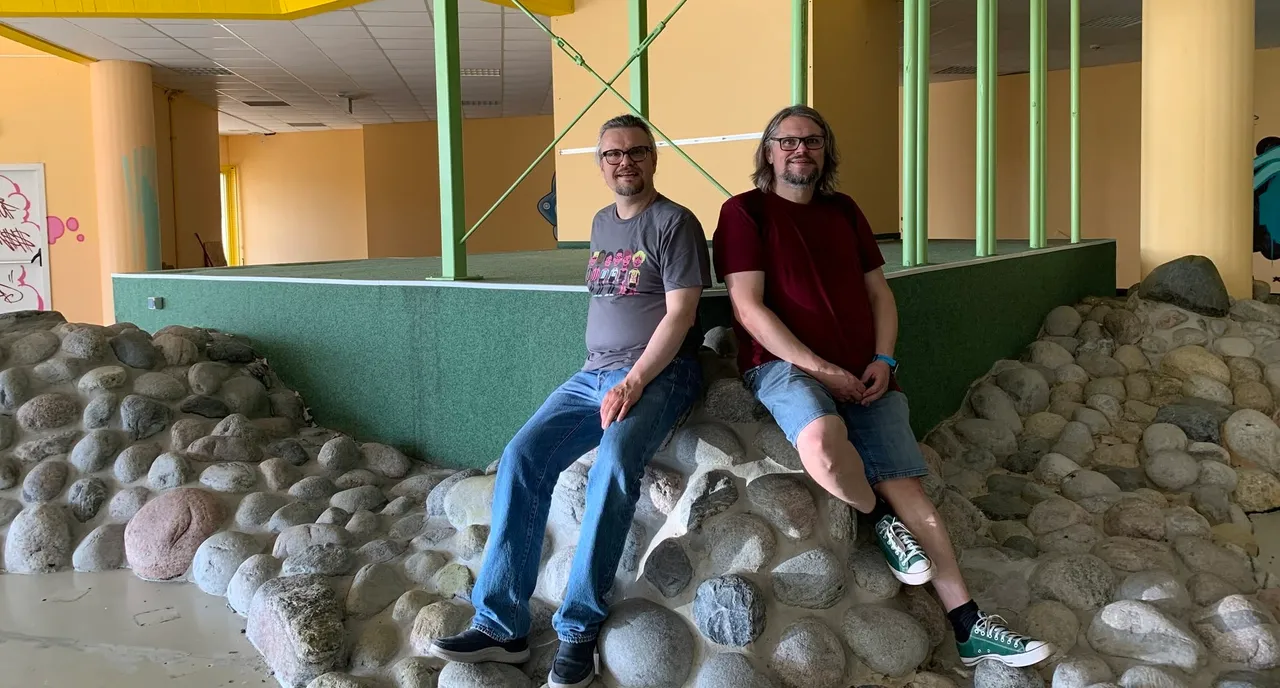 Sebastian Göttling und Simon Fistrich sitzen auf einem Steinhaufen in einem Raum mit gelben Wänden und grünen Säulen.