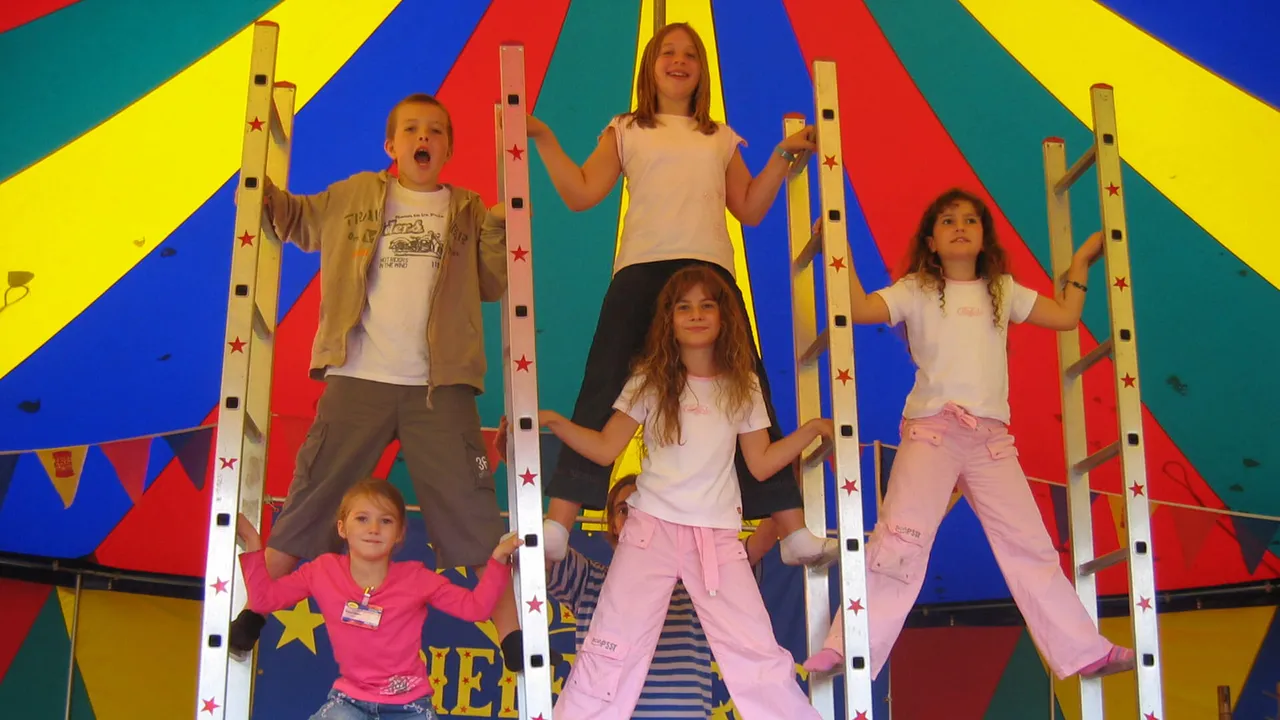 Kinder des Kölner Spielezirkus posieren auf Leitern unter einem bunten, gestreiften Zelt.