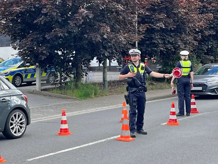 Polizisten regeln den Verkehr an einer Straßensperre mit Warnwesten, Absperrband und Verkehrskegeln. Im Hintergrund sind weitere Einsatzfahrzeuge und Autos zu sehen.