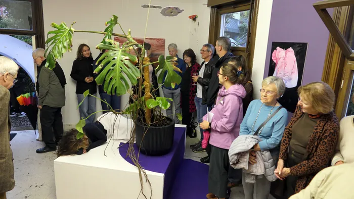 Gäste der Vernissage „Zeitmangelwesen“ betrachten eine Installation mit einer großen Monstera-Pflanze und einer auf einem Podest liegenden Person.
