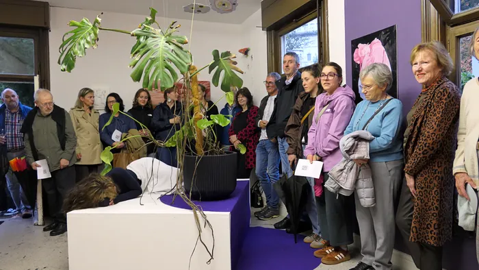 Besucher der Vernissage der Ausstellung „Zeitmangelwesen“ stehen um eine große Monstera-Pflanze auf einem Podest, an dem eine Person lehnt.