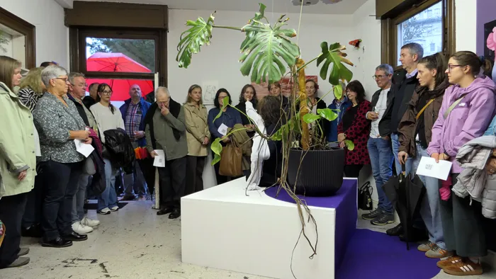 Eine Gruppe von Menschen steht bei der Vernissage der Ausstellung „Zeitmangelwesen“ um eine große Monstera-Pflanze auf einem weißen Podest.
