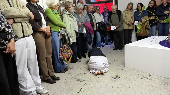 Eine Person in weißer Kleidung liegt bäuchlings auf dem Boden, während eine Gruppe von Menschen sie in einem Ausstellungsraum beobachtet.