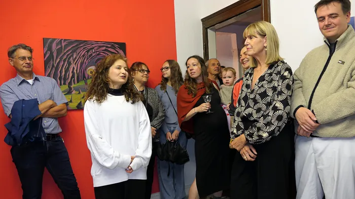 Eine Gruppe von Gästen bei der Vernissage der Ausstellung „Zeitmangelwesen“ vor einer roten Wand mit einem abstrakten Gemälde.