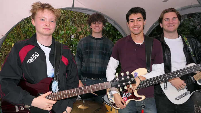 Vier junge Männer mit Gitarren und einem Bass stehen vor einer grünen Hecke unter einem weißen Zeltdach.