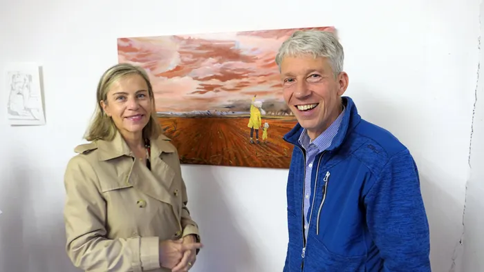 Eine Frau und ein Mann lächeln vor einem Gemälde einer Landschaft mit einer Person in gelber Kleidung.
