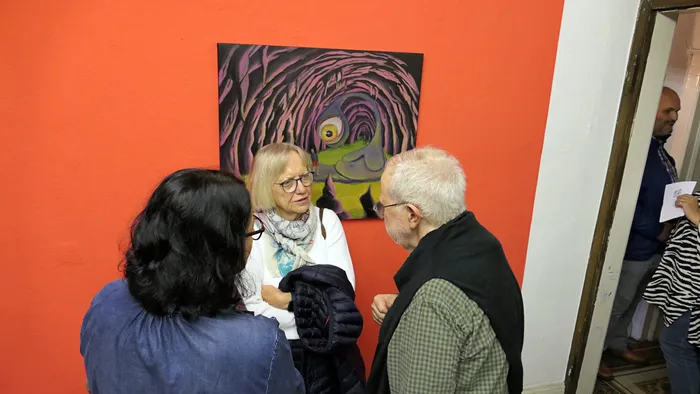 Drei Personen im Gespräch vor einem abstrakten Gemälde an einer orangefarbenen Wand während der Vernissage der Ausstellung „Zeitmangelwesen“.