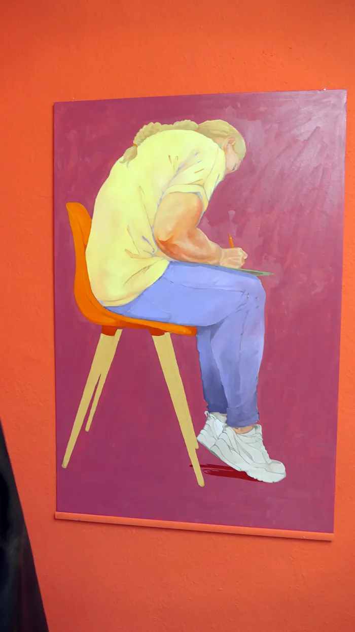 Gemälde einer Person in gelbem Oberteil und blauer Hose, die vornübergebeugt auf einem orangefarbenen Stuhl schreibt.