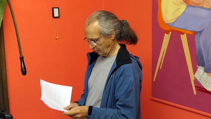 Ein Mann mit Brille und langem Zopf liest ein weißes Blatt Papier vor einer orangefarbenen Wand.