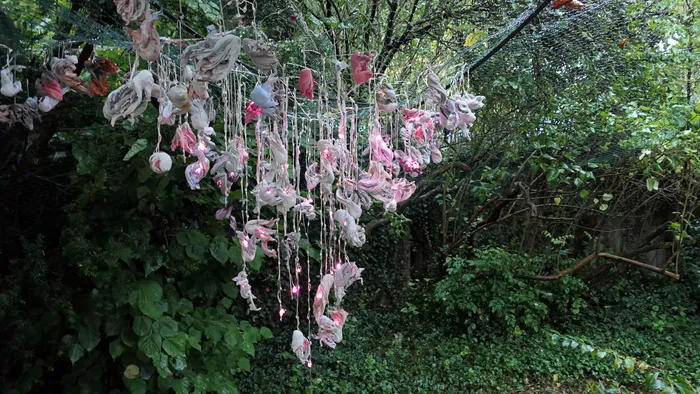 Hängende Textilinstallation mit rosa beleuchteten Stoffelementen vor dichtem grünem Blattwerk.