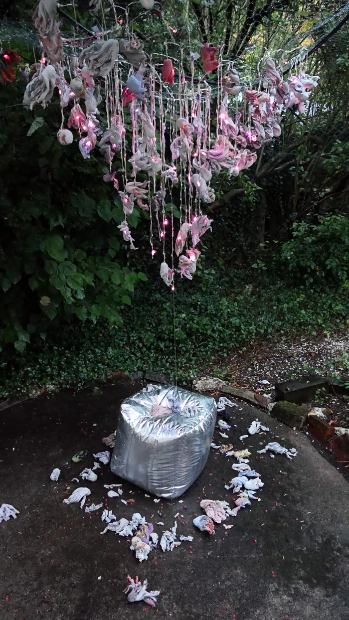 Kunstinstallation mit hängenden, beleuchteten Stoffelementen in Rosa und Weiß über einem großen silbernen Bündel auf dem Boden.