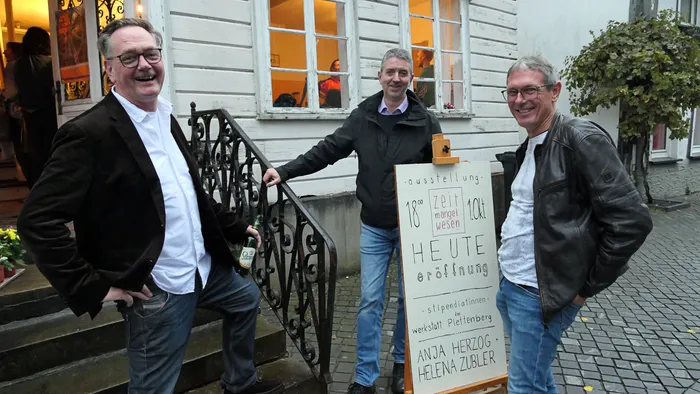 Drei Männer stehen lächelnd vor einem Schild zur Eröffnung der Ausstellung „Zeitmangelwesen“ von Anja Herzog und Helena Zubler.