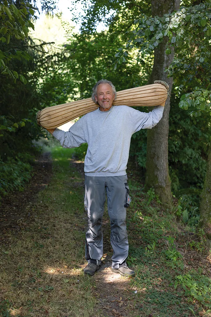 Günter Wermkes steht auf einem Waldweg und trägt ein großes, geripptes, längliches Objekt auf seinen Schultern.