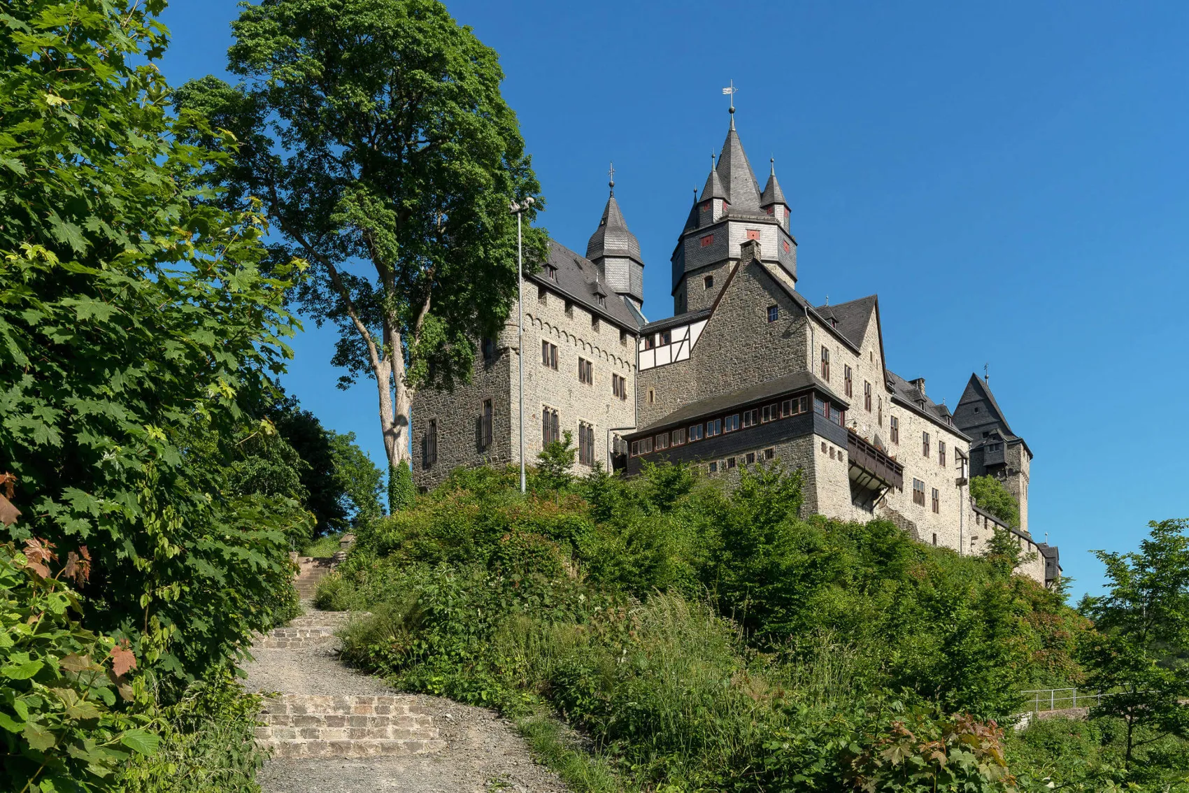 Graue Burg auf einem bewachsenen Hügel mit einem Pfad, der zur Burg führt. Im Hintergrund blauer Himmel.