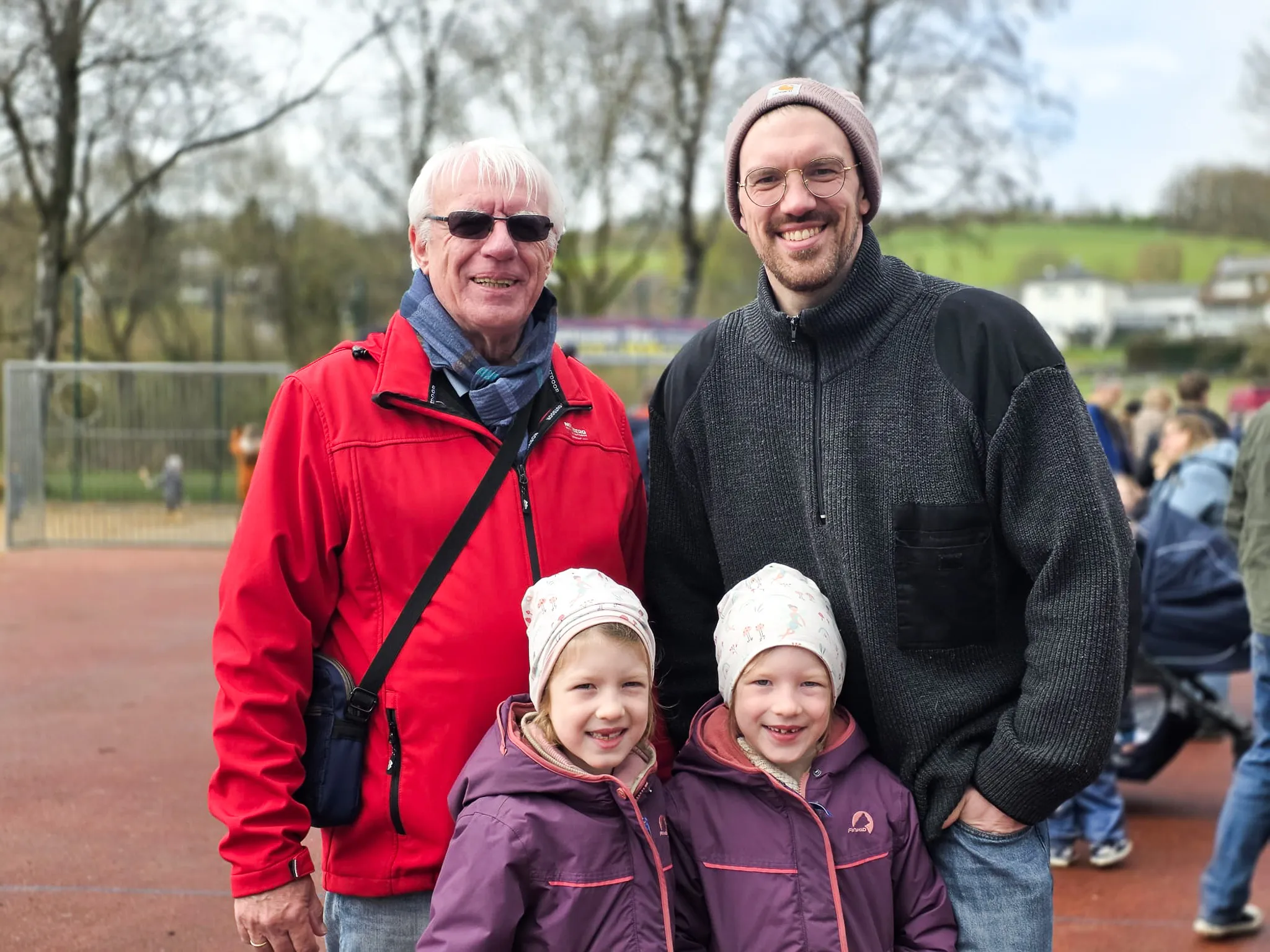 Zwei Generationen lächeln in die Kamera: Ein älterer Mann in roter Jacke und ein jüngerer Mann mit Bart stehen mit zwei kleinen Mädchen in Wintermützen vor einer roten Sportfläche.
