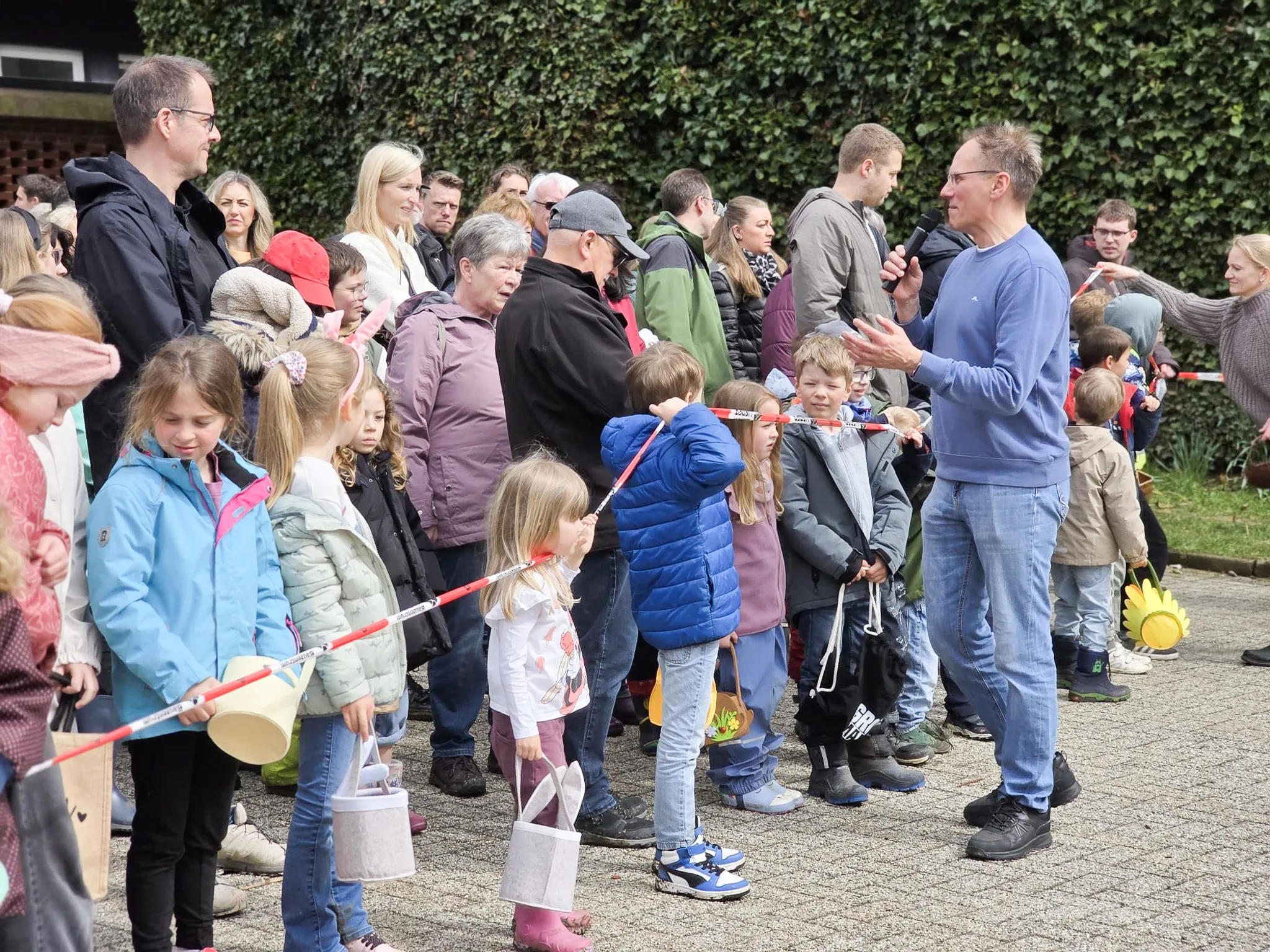 Eine Gruppe von Erwachsenen und Kindern steht an einer abgesperrten Fläche, während ein Mann gestikulierend etwas erklärt. Im Hintergrund sind weitere Menschen zu sehen, einige halten Eimer oder Körbe bereit.