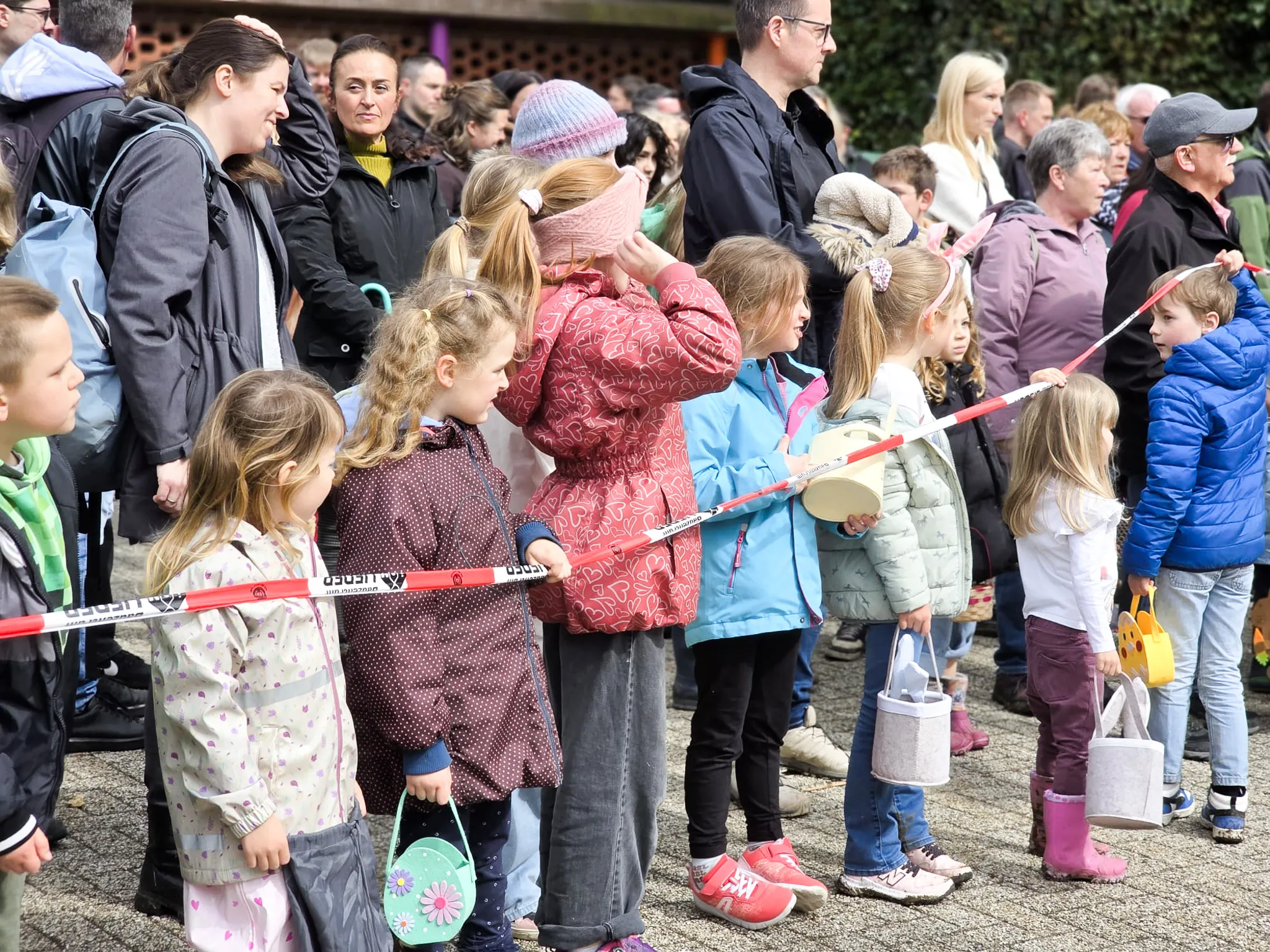 Kinder stehen hinter einer roten Absperrkordel und warten auf den Start der Ostereiersuche, umgeben von Eltern und Zuschauern.