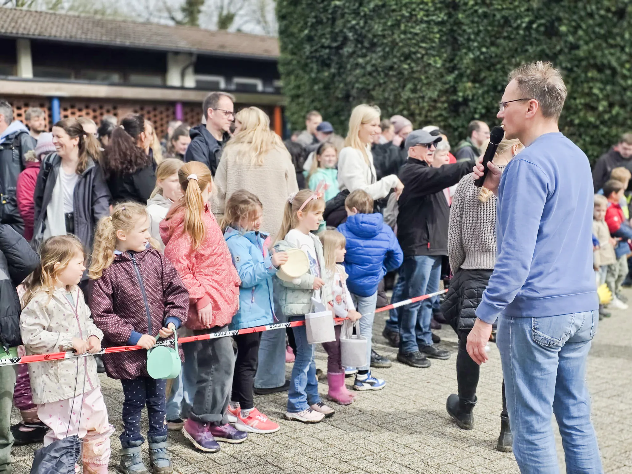 Eine Schlange von Kindern mit Eimern steht vor einem Mann mit Mikrofon, während im Hintergrund weitere Familien stehen. Die Ostereiersuche findet im Freien statt.