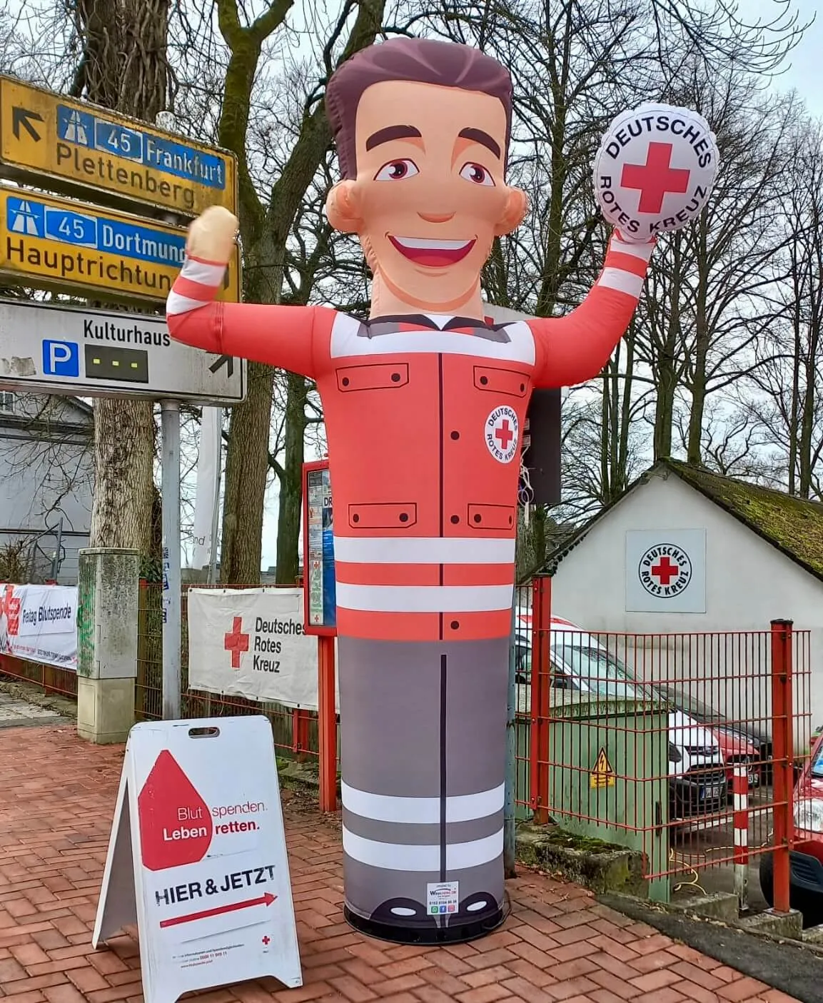 Aufblasbare Figur des Deutschen Roten Kreuzes winkt vor einem Ortsschild und einem Plakat zur Blutspendeaktion.