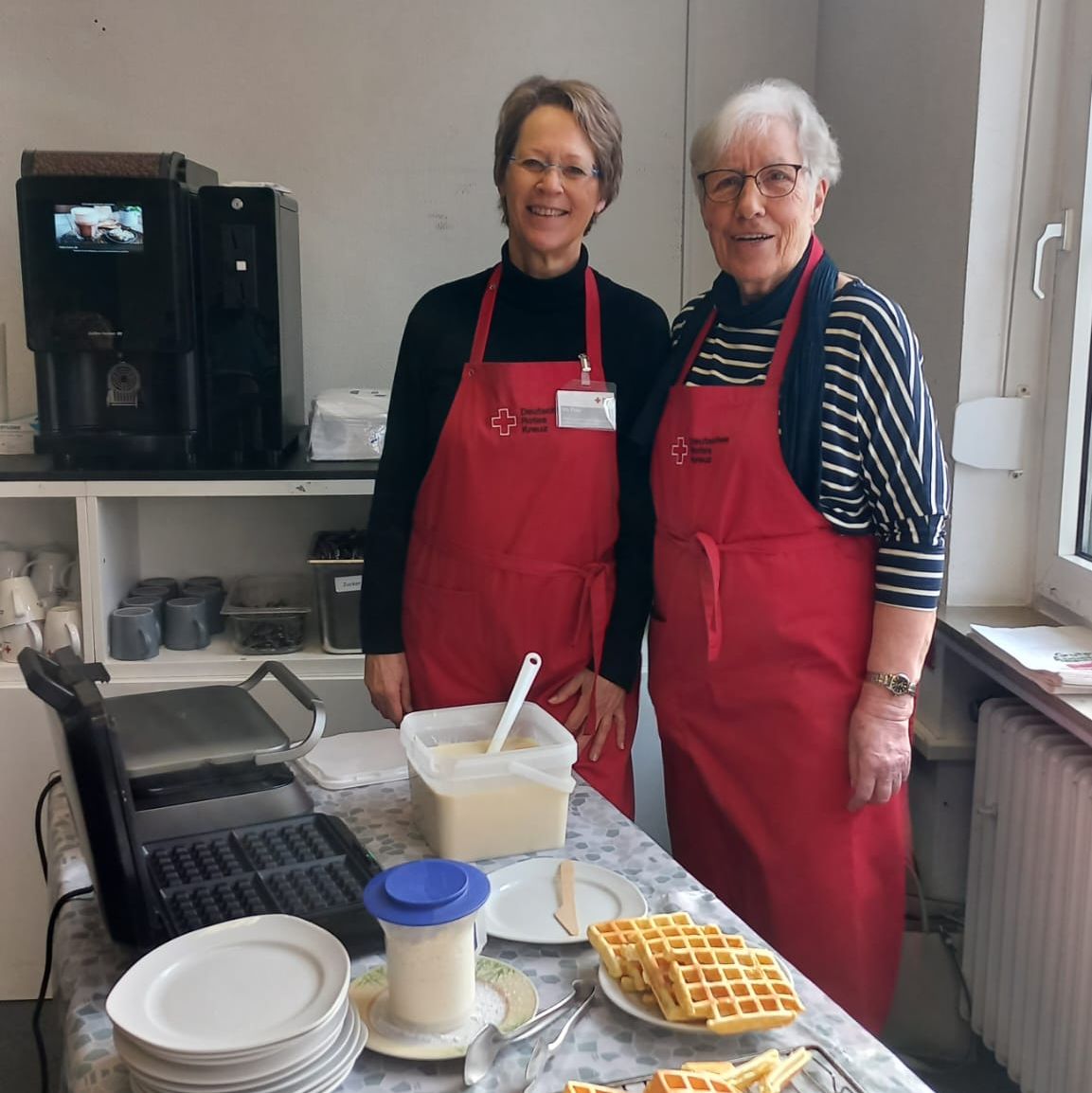 Zwei Frauen mit roten Schürzen und dem Logo des Deutschen Roten Kreuzes stehen an einem Tisch mit Waffeln und Geschirr.