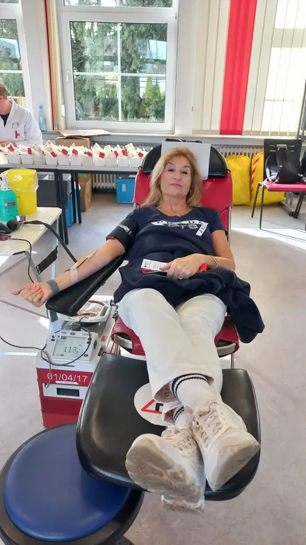 Eine Frau liegt während einer Blutspende in einem roten Sessel. An ihrem Arm ist ein Blutdruckmessgerät angebracht und eine Blutentnahme läuft. Im Hintergrund sind weitere Spender und medizinische Geräte zu sehen.
