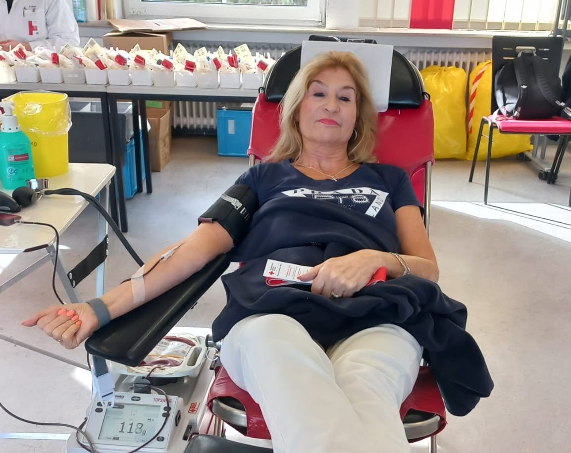 Eine Frau liegt während einer Blutspende in einem roten Sessel. An ihrem Arm ist ein Blutdruckmanschette angelegt und eine Blutentnahme läuft.