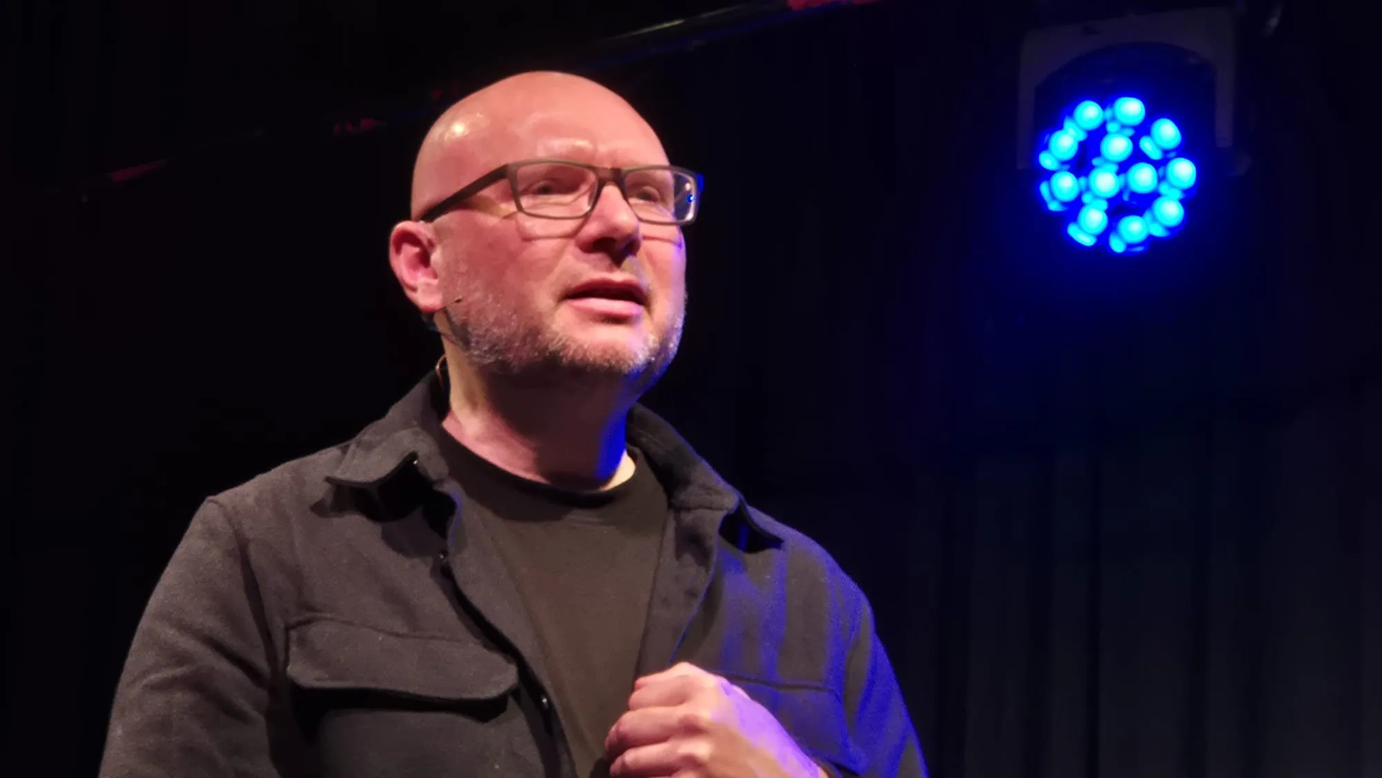 Ein Mann mit Glatze und Brille spricht in einen Raum. Im Hintergrund ist ein blaues Licht zu sehen.