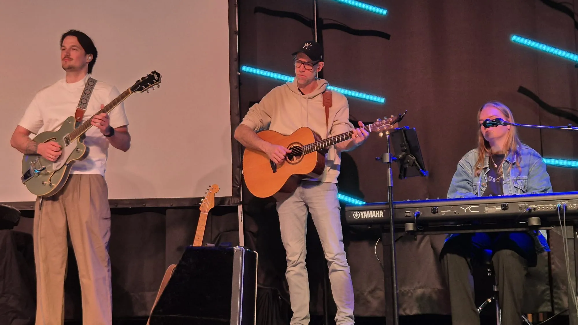Drei Musiker spielen auf einer Bühne: Ein Mann mit einer E-Gitarre links, ein Mann mit einer Akustikgitarre in der Mitte und eine Frau mit einem Keyboard rechts. Im Hintergrund eine Leinwand und blaue Lichtelemente.