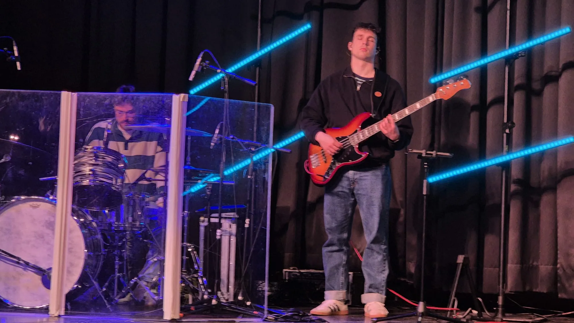 Ein Gitarrist spielt vor einer transparenten Plexiglaswand, hinter der ein Schlagzeuger sitzt. Im Hintergrund sind blaue Lichtstreifen zu sehen.