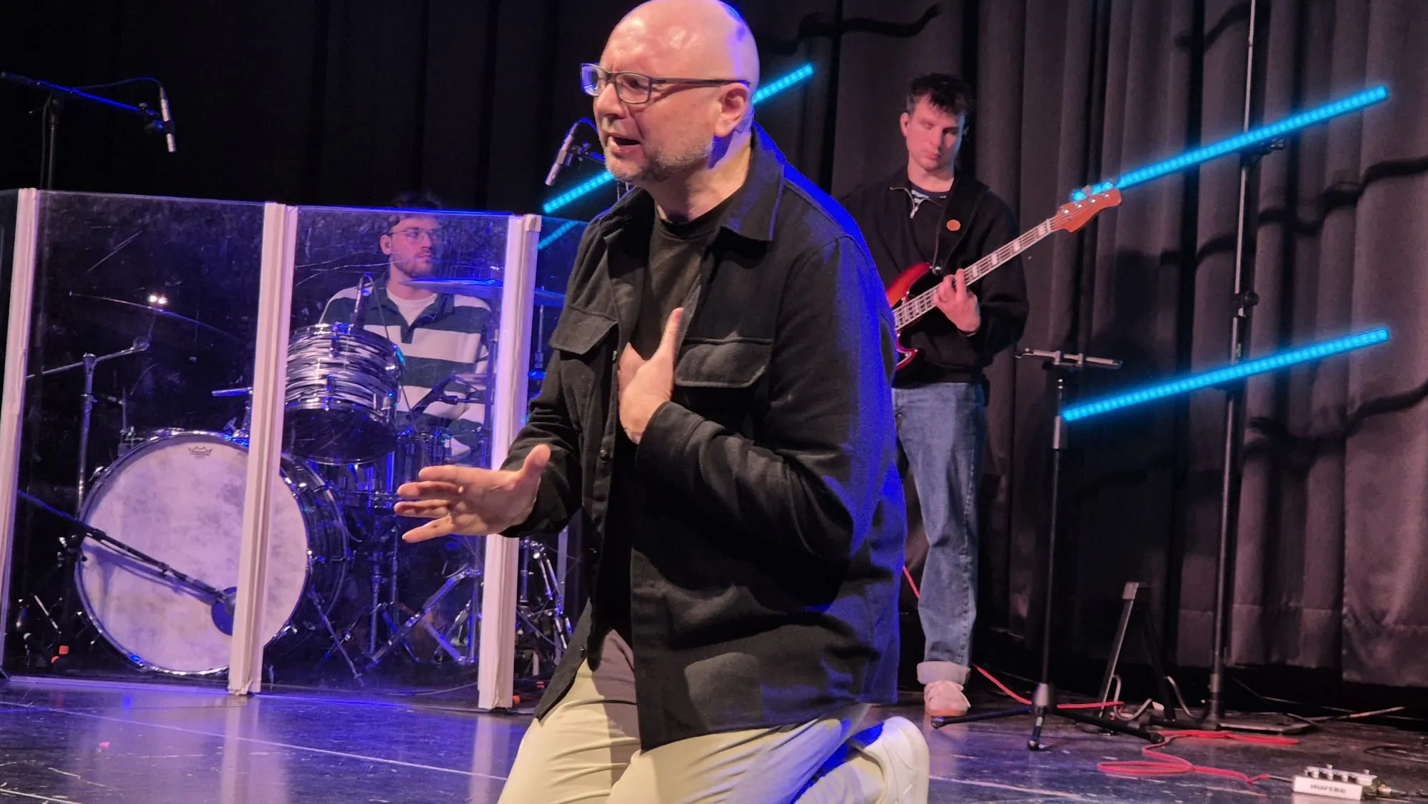 Ein Mann mit Glatze und Brille gestikuliert während eines Auftritts auf einer Bühne. Im Hintergrund sind ein Schlagzeuger und ein Bassist zu sehen.