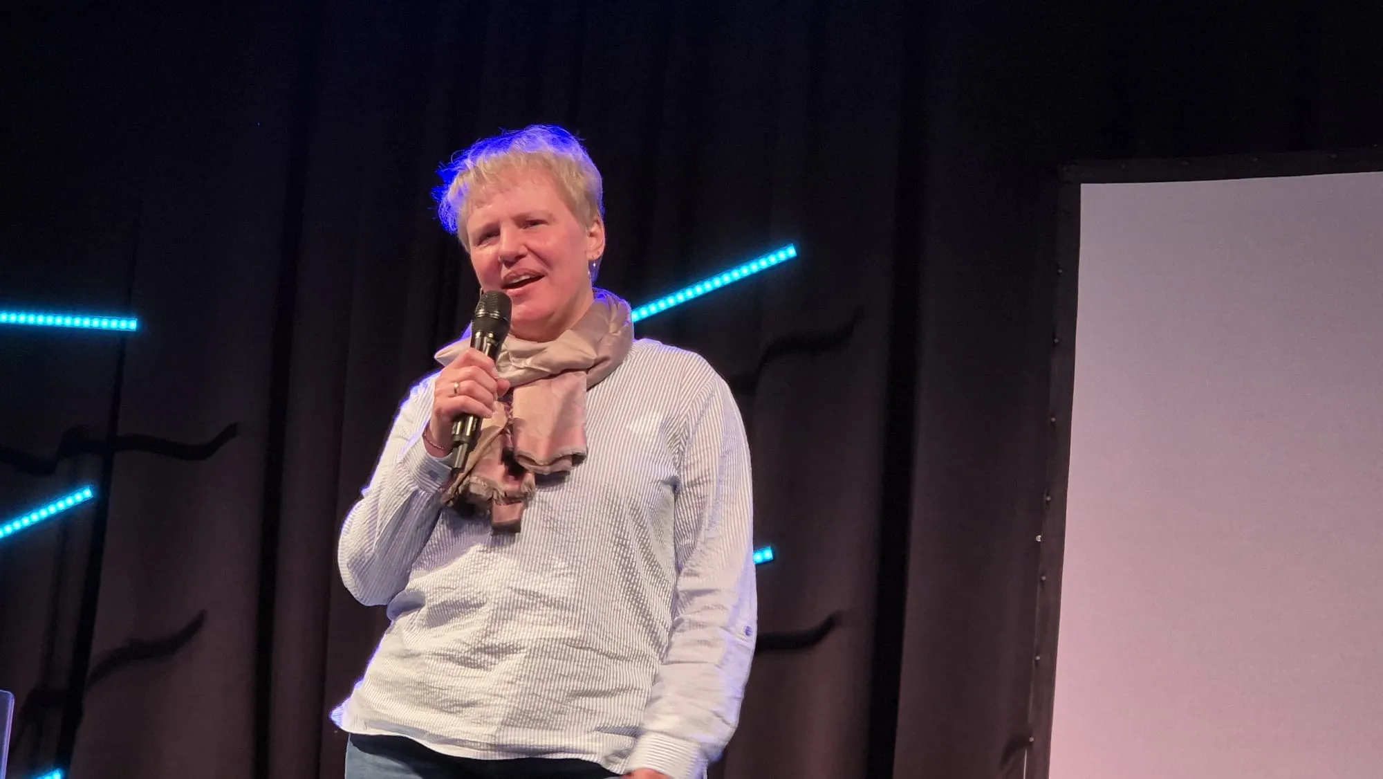 Frau mit kurzem, grauem Haar hält ein Mikrofon und spricht auf einer Bühne vor Publikum.
