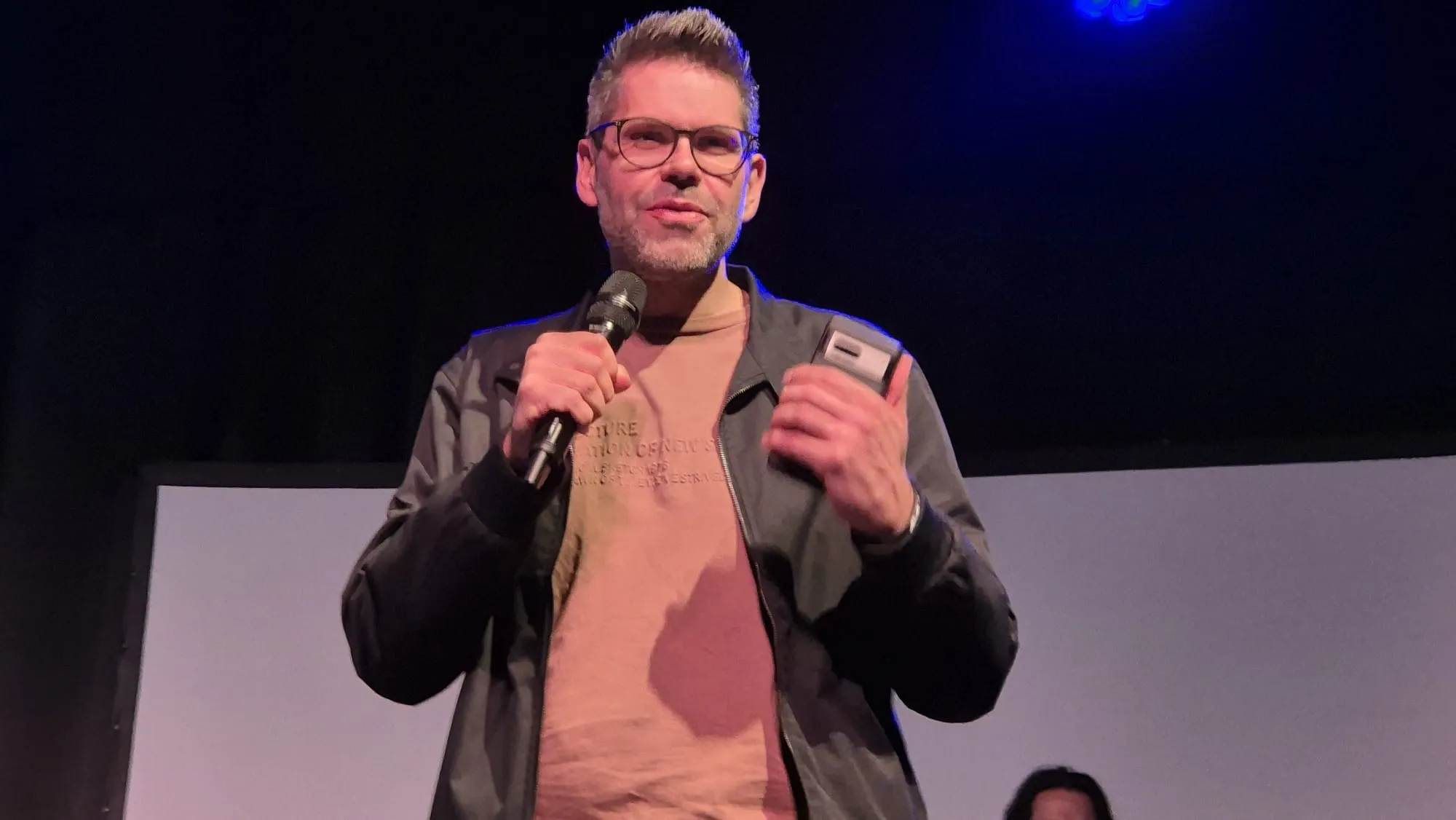 Mann mit Brille und Mikrofon hält eine Rede vor Publikum in einem dunklen Raum.