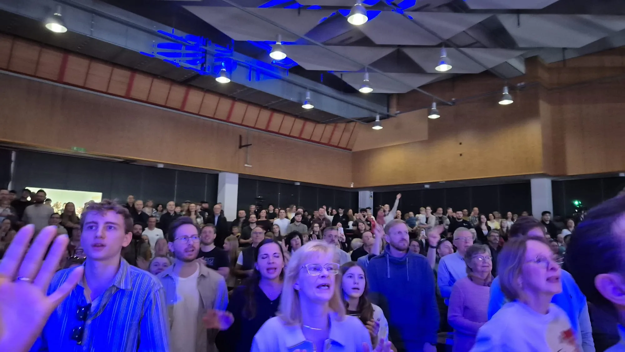 Große Menschenmenge in einem hellen Raum mit einer hohen Decke und vielen Deckenleuchten. Die Menschen blicken nach oben, einige heben die Hände.