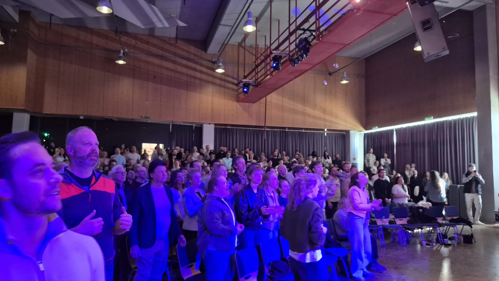 Große Menschenmenge in einem hellen Saal mit Holzwänden und einer Bühne. Viele Menschen sitzen auf Stühlen und schauen nach vorne.