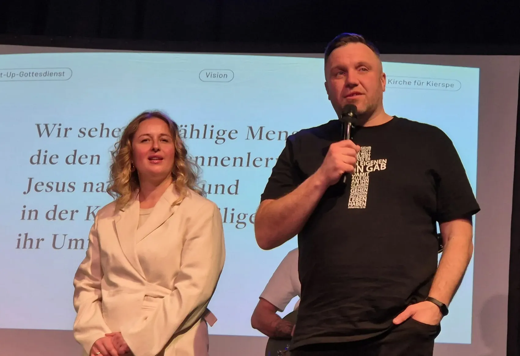 Andreas und Nadiya Matievsky stehen auf einer Bühne vor einer Projektion mit Text und sprechen vor einem Publikum. Andreas hält ein Mikrofon.