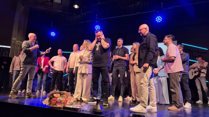 Eine Gruppe von etwa zwanzig Personen steht auf einer Bühne vor einem dunklen Hintergrund. Im Vordergrund hält ein Mann mit Bart und schwarzem T-Shirt einen Blumenstrauß und scheint emotional zu sein.