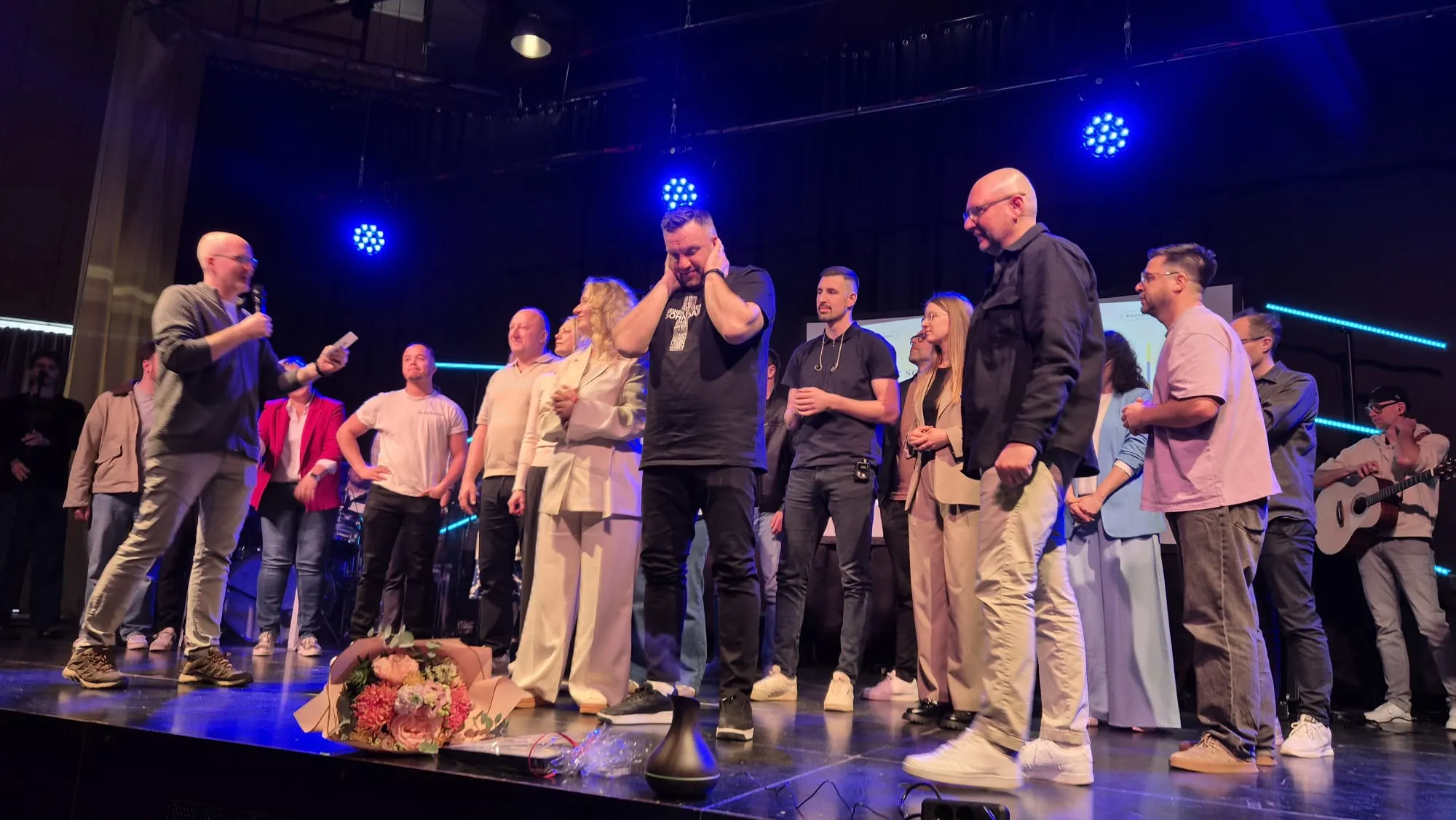 Eine Gruppe von etwa zwanzig Personen steht auf einer Bühne vor einem dunklen Hintergrund. Im Vordergrund hält ein Mann mit Bart und schwarzem T-Shirt einen Blumenstrauß und scheint emotional zu sein.