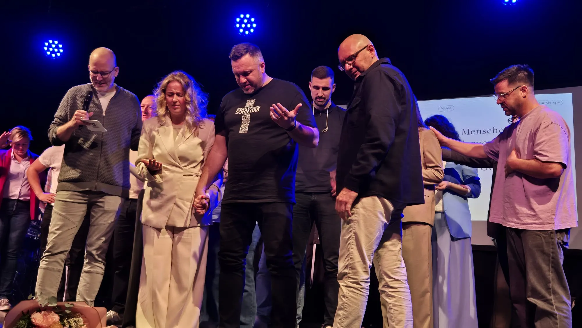 Mehrere Personen stehen auf einer Bühne und halten Hände, während ein Mann mit einem Tablet spricht. Im Hintergrund ist eine Präsentation mit dem Wort „Menschen“ zu sehen.