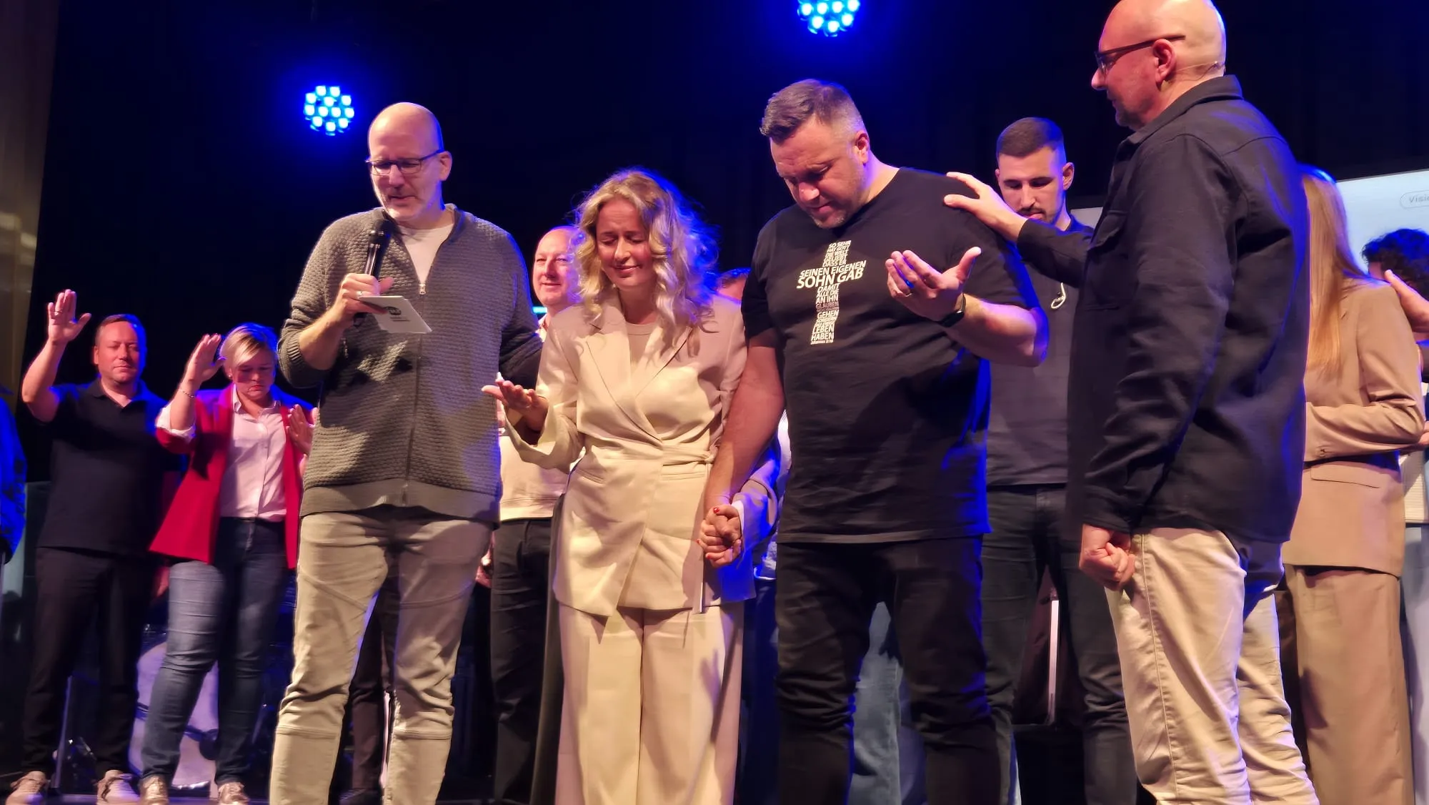 Eine Gruppe von etwa zehn Menschen steht auf einer Bühne und hält Hände, während sie nach oben blickt. Im Hintergrund weitere Personen und eine große Lichtinstallation.