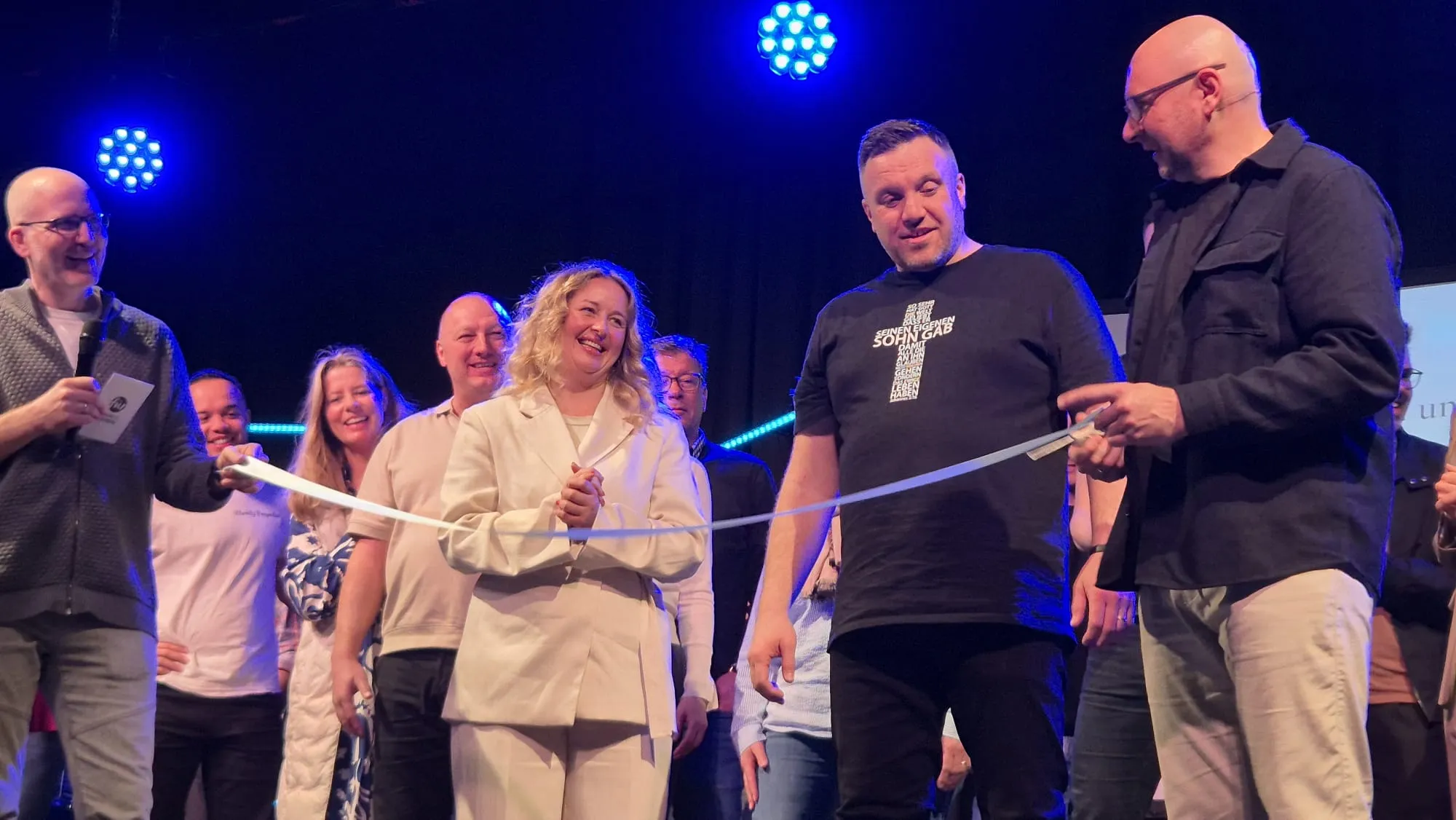 Zwei Männer schneiden ein weißes Band mit einer Schere durch, umgeben von einer Gruppe lächelnder Menschen bei einer Eröffnungsfeier.