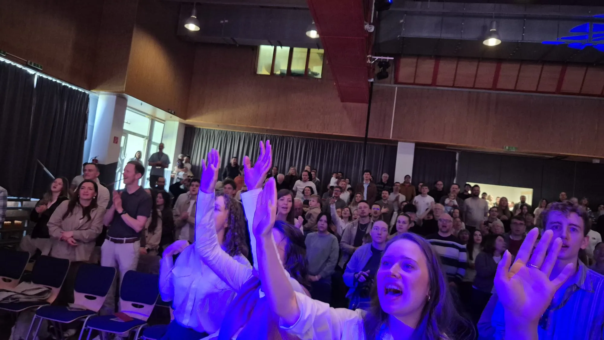 Große Menschenmenge in einem hellen Saal mit erhöhter Bühne, viele heben die Hände. Im Hintergrund eine Empore und ein rotes Element an der Decke.
