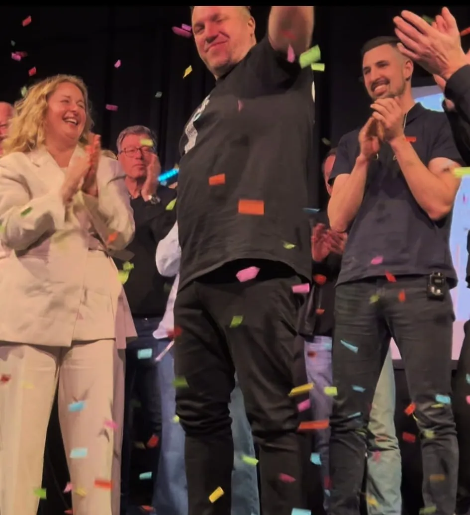 Ein lächelnder Mann im schwarzen Shirt steht im Mittelpunkt auf einer Bühne, umgeben von applaudierenden Menschen und Konfetti. Im Hintergrund weitere Personen und ein dunkler Vorhang.